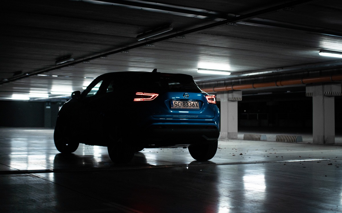 Niebieski samochód osobowy, tyłem, z włączonymi światłami pozycyjnymi, zaparkowany w ciemnym garażu podziemnym z betonową podłogą.