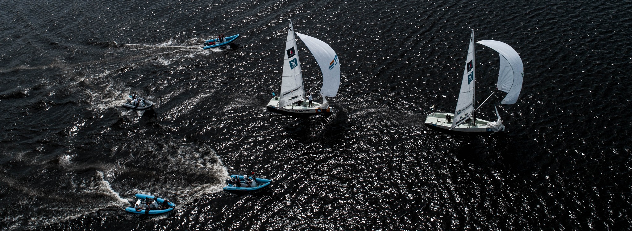 Widok z góry na regaty żeglarskie: dwie żaglówki z białymi żaglami i trzy motorówki na ciemnej, wzburzonej wodzie.