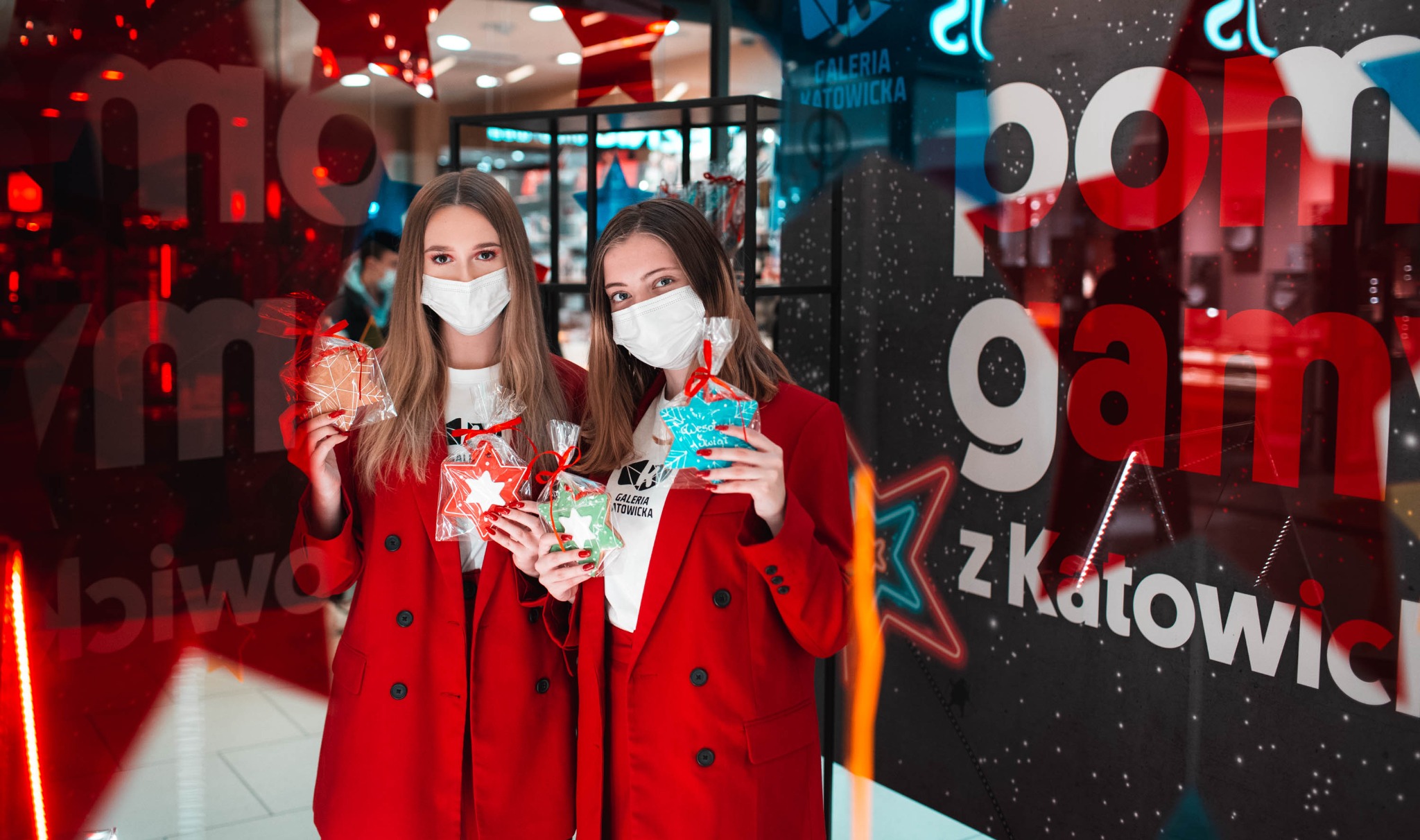 Dwie kobiety w czerwonych marynarkach i maseczkach ochronnych trzymają świąteczne pierniki w kształcie gwiazdek przed witryną sklepową Galerii Katowickiej z neonowymi dekoracjami świątecznymi.