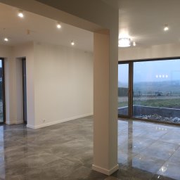 PrimaSort Remont - Nowoczesne wnętrze z szarą marmurową podłogą, dużymi oknami z widokiem na zewnątrz i punktowym oświetleniem sufitowym, widoczna kolumna konstrukcyjna.