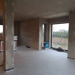 PrimaSort Remont - Surowy stan wnętrza w trakcie prac: ściany po szpachlowaniu, widoczne kable zwisające z sufitu, podłoga pokryta płytami izolacyjnymi, okna z widokiem na zewnątrz, materiały budowlane Besser w tle.