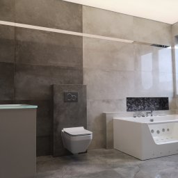 PrimaSort Remont - Nowoczesna łazienka z wanną z hydromasażem, podwieszaną toaletą i umywalką z szarą szafką, wykończona płytkami w odcieniach szarości i beżu.