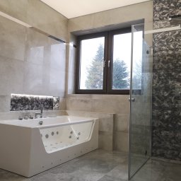PrimaSort Remont - Nowoczesna łazienka z dużą, białą wanną z hydromasażem, przeszkloną kabiną prysznicową i szarą, geometryczną mozaiką na ścianie prysznica, widok na drzewa za oknem.