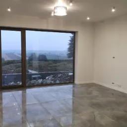 Pusty pokój z dużym oknem tarasowym, szare płytki na podłodze, białe ściany i sufit z oświetleniem punktowym oraz ozdobną lampą wiszącą; widok z okna na krajobraz po deszczu.