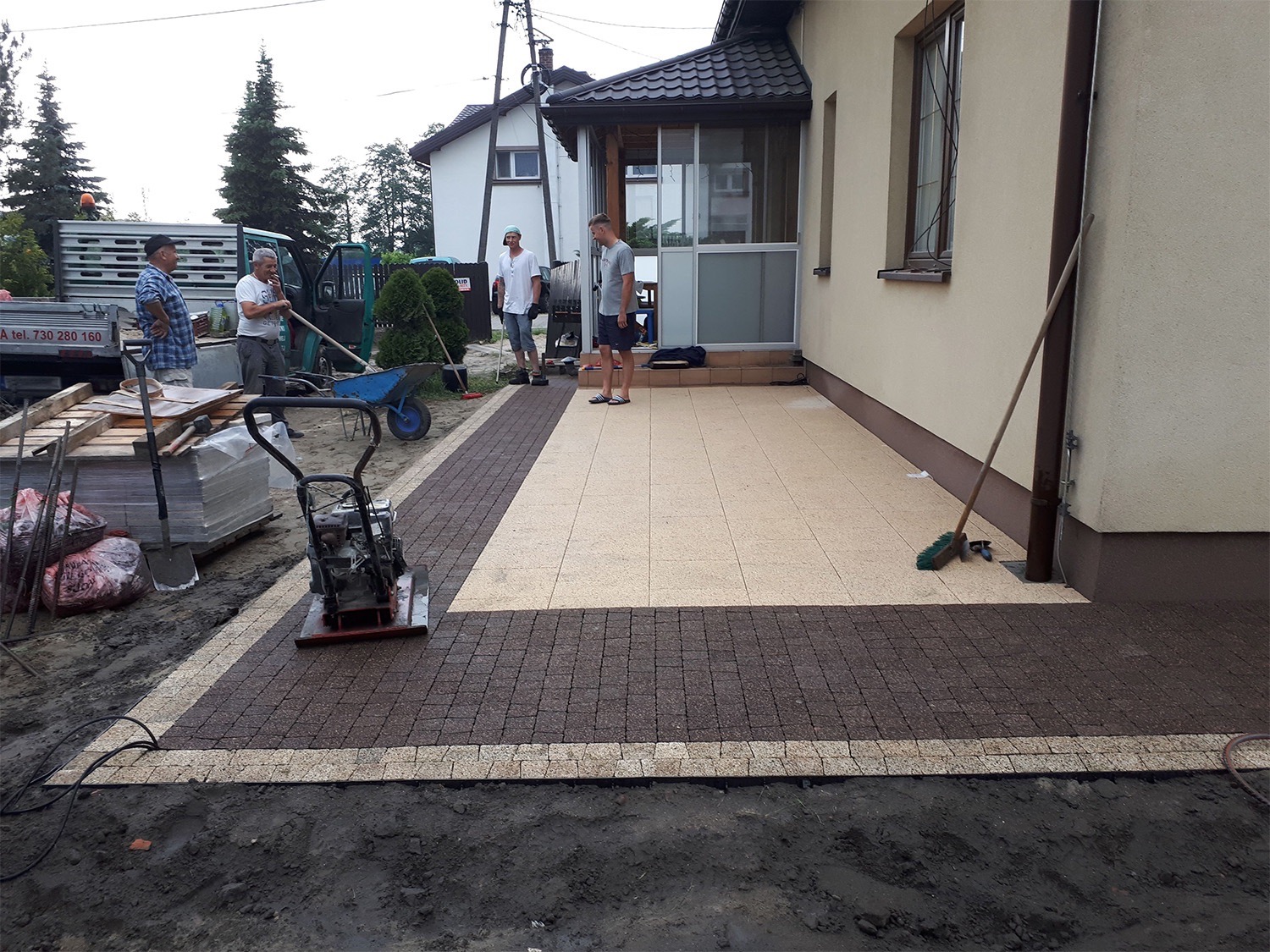 Układanie kostki brukowej na tarasie przy domu; w tle ekipa budowlana z ubijarką wibracyjną, widoczne narzędzia i materiały budowlane.