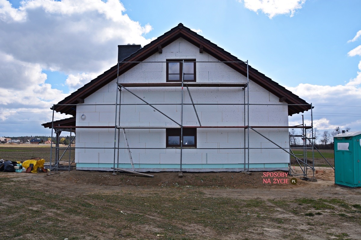 Dom w trakcie ocieplania styropianem, widoczne rusztowania, okna z brązowymi ramami oraz toaleta przenośna na zewnątrz.