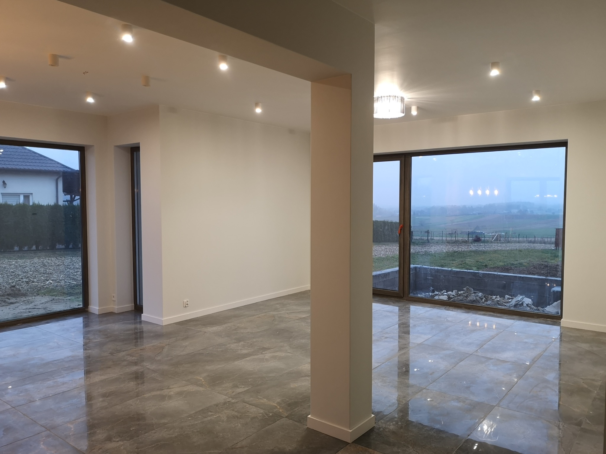 Nowoczesne wnętrze z szarą marmurową podłogą, dużymi oknami z widokiem na zewnątrz i punktowym oświetleniem sufitowym, widoczna kolumna konstrukcyjna.