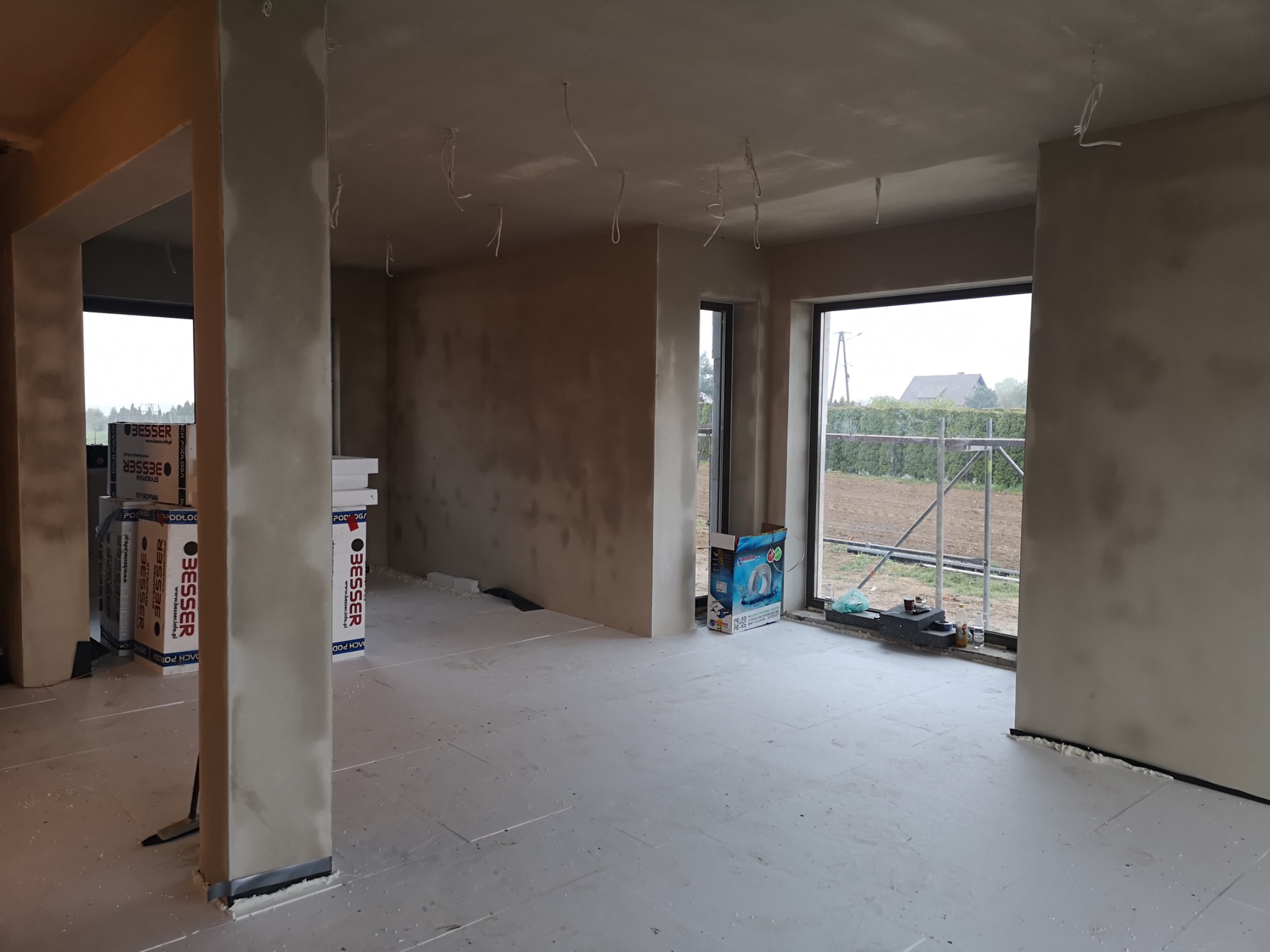 Surowy stan wnętrza w trakcie prac: ściany po szpachlowaniu, widoczne kable zwisające z sufitu, podłoga pokryta płytami izolacyjnymi, okna z widokiem na zewnątrz, materiały budowlane Besser w tle.