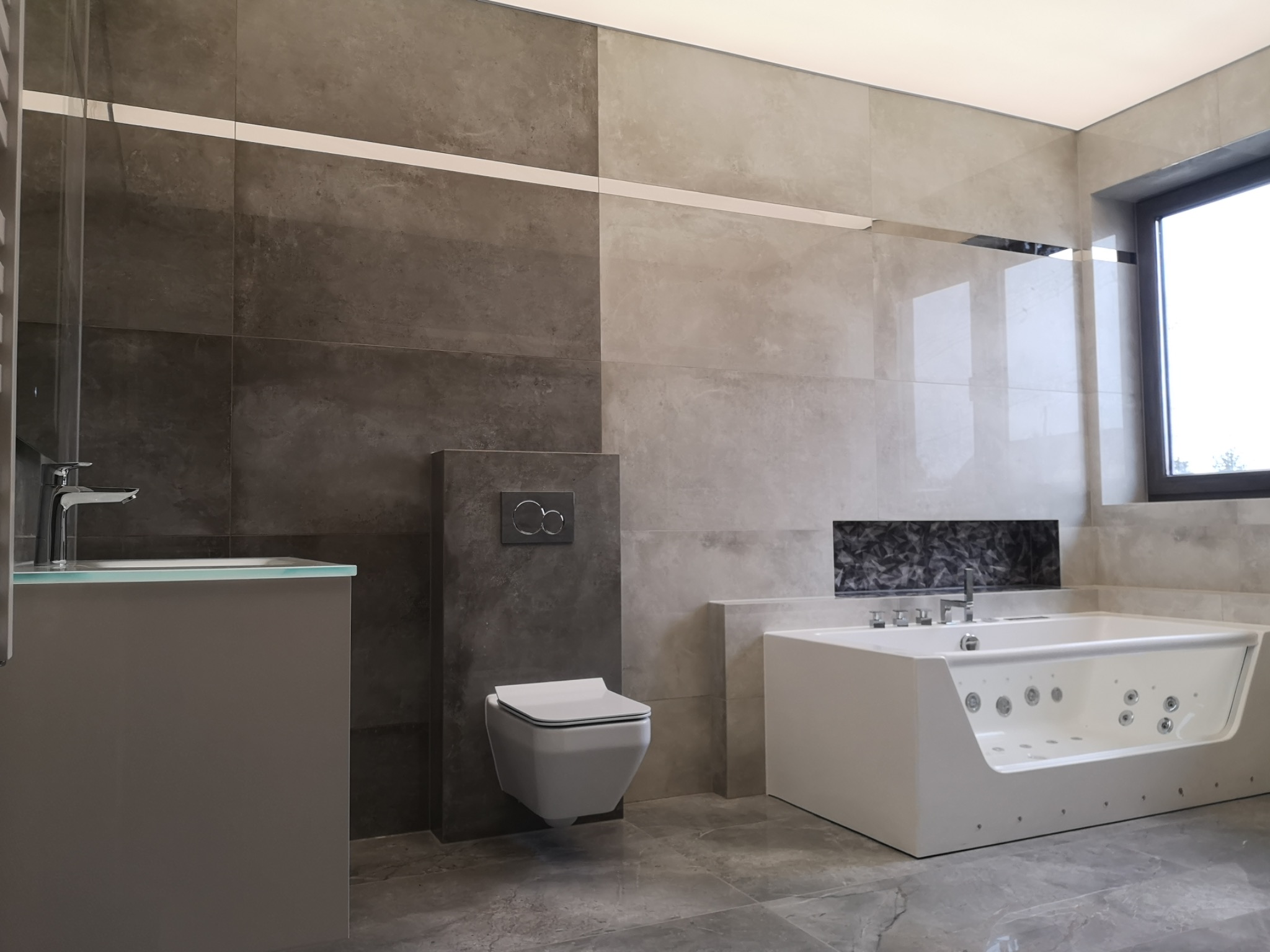 Nowoczesna łazienka z wanną z hydromasażem, podwieszaną toaletą i umywalką z szarą szafką, wykończona płytkami w odcieniach szarości i beżu.