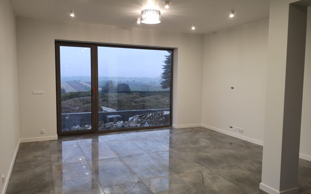 Pusty pokój z dużym oknem tarasowym, szare płytki na podłodze, białe ściany i sufit z oświetleniem punktowym oraz ozdobną lampą wiszącą; widok z okna na krajobraz po deszczu.