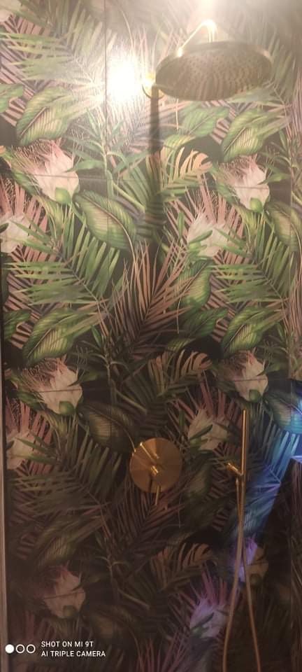 Kabina prysznicowa z deszczownicą i złotą armaturą na tle ściany wyłożonej płytkami z motywem tropikalnych liści.