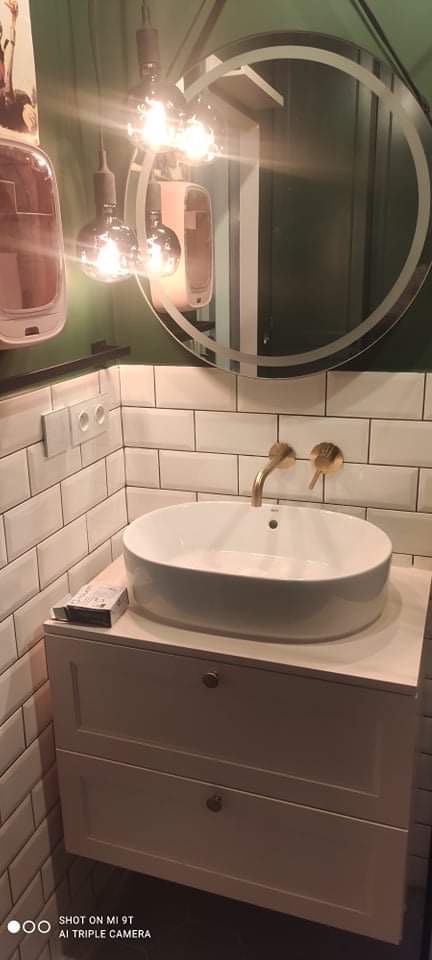 Biała umywalka nablatowa o owalnym kształcie z baterią w kolorze szczotkowanego złota, zamontowana na białej szafce z dwiema szufladami, obok widoczna paczka chusteczek, ściana wyłożona białymi...