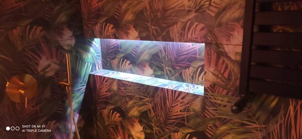 Nowoczesna łazienka z tropikalną tapetą, podświetlaną wnęką i złotą armaturą.