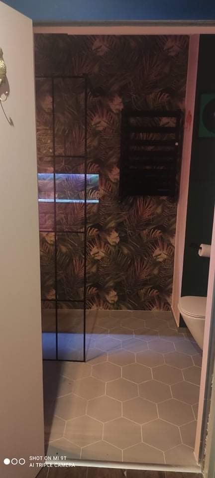 Nowoczesna łazienka z heksagonalnymi płytkami podłogowymi, tropikalną tapetą, czarną kabiną prysznicową typu walk-in i podświetlaną wnęką.