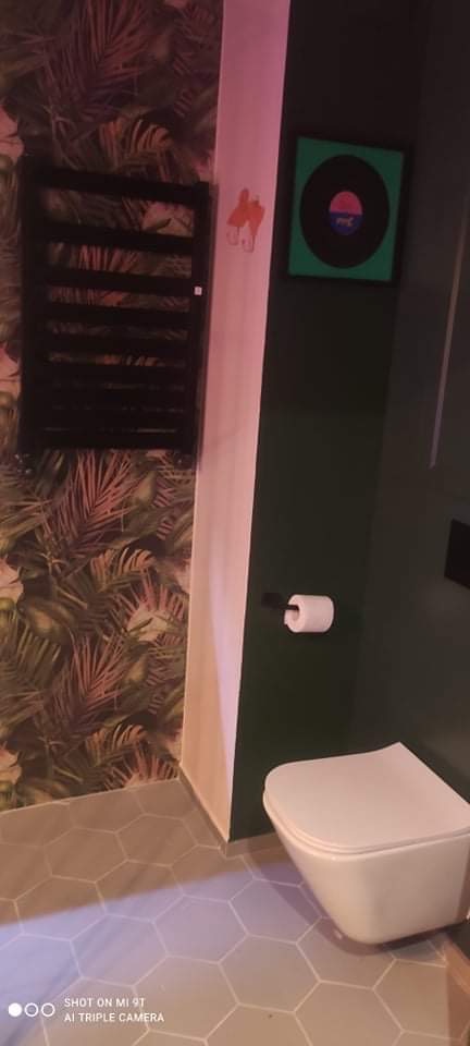 Nowoczesna łazienka z heksagonalną szarą podłogą, toaleta podwieszana, ciemnozielona ściana z uchwytem na papier toaletowy, czarny grzejnik drabinkowy na ścianie w tropikalne liście i abstrakcyjny...