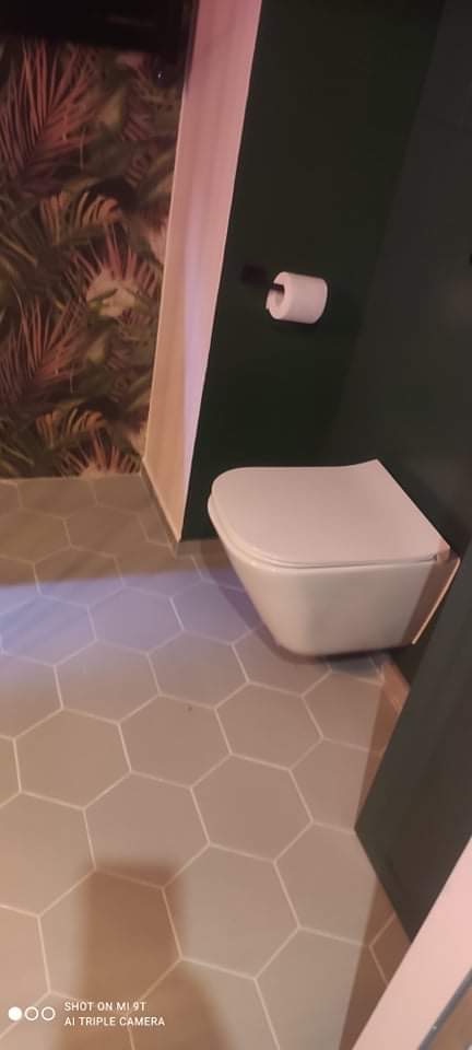 Wisząca toaleta z deską wolnoopadającą na tle ciemnozielonej ściany i heksagonalnej podłogi w odcieniach beżu, obok ściana z tapetą w motywy tropikalne.