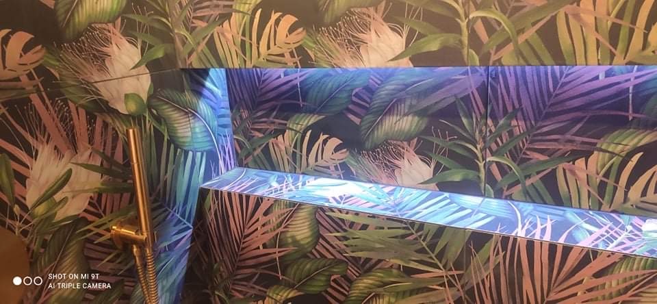 Ściana łazienki wykończona płytkami z motywem tropikalnych liści, podświetlona niebieskim światłem LED, z widoczną złotą baterią prysznicową.