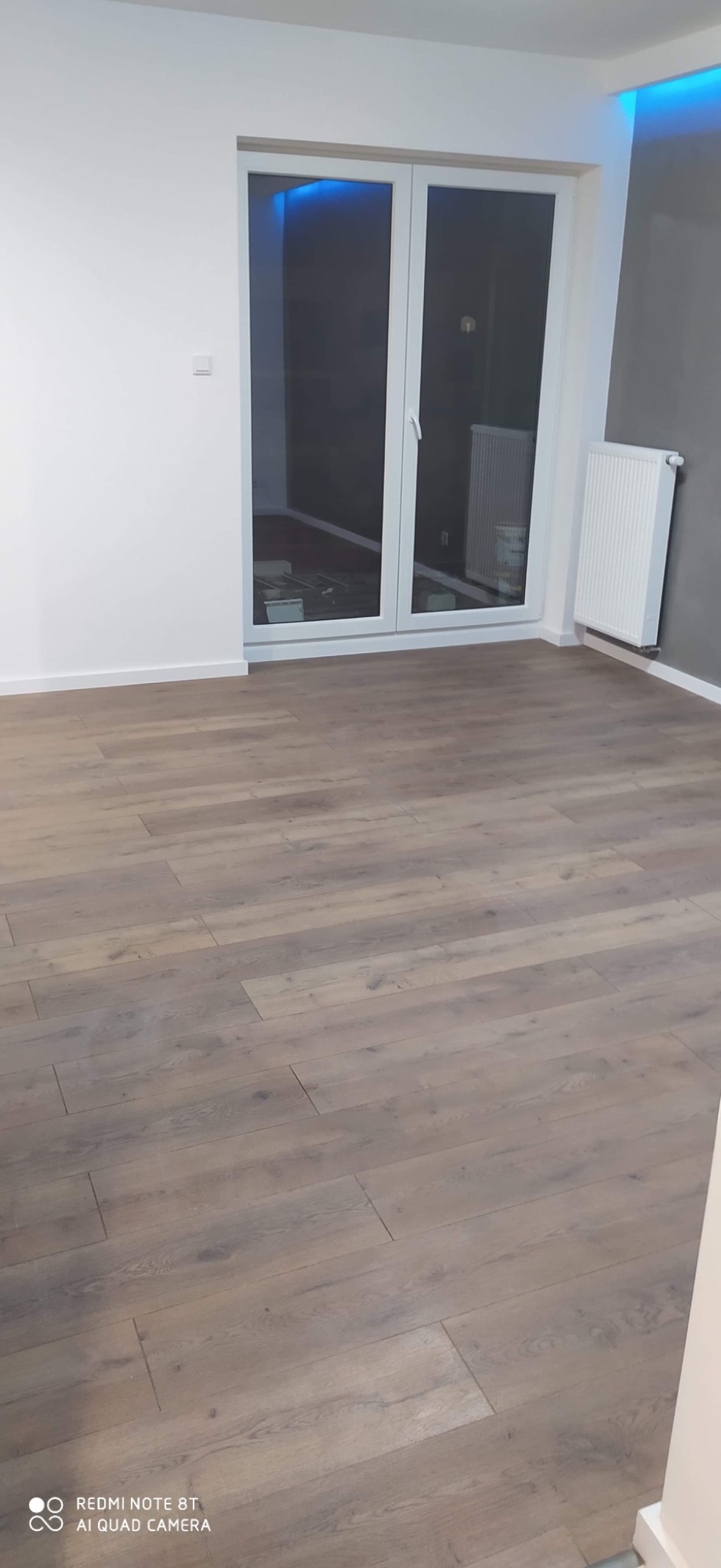 Ułożona podłoga z paneli imitujących drewno w nowym pokoju, widoczne białe ściany, grzejnik i okno balkonowe z podświetleniem LED w górnej części ramy.