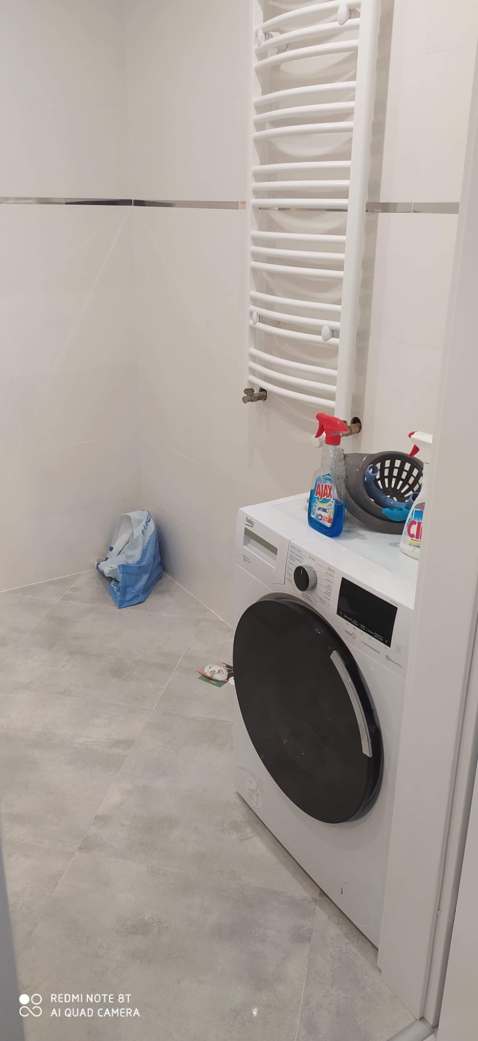 Wykończona łazienka z białymi płytkami z dekoracyjnymi paskami, grzejnikiem drabinkowym, pralką Beko i środkami czystości na pralce.