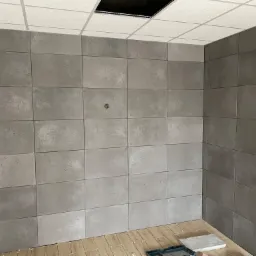 Ściany w pomieszczeniu wyłożone szarymi, betonowymi płytkami imitującymi panele, z widocznymi punktami mocowania, na podłodze widoczne fragmenty drewnopodobnych płytek oraz narzędzia glazurnicze.