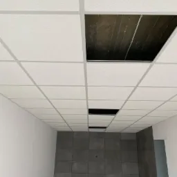 Sufit podwieszany kasetonowy z widocznymi otworami instalacyjnymi i fragmentem ściany z szarymi płytkami ceramicznymi