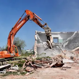 Pomarańczowa koparka Doosan z hydraulicznymi nożycami kruszy resztki ceglanego budynku, tumany kurzu w słoneczny dzień.