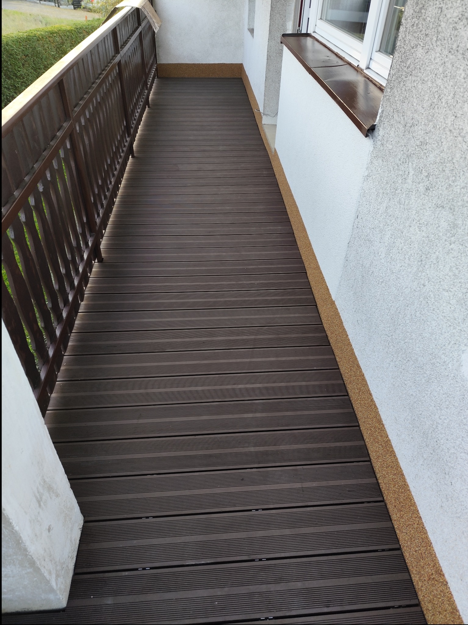 Wykończony balkon z ciemnobrązową podłogą z desek kompozytowych, widoczne balustrady i fragment ściany budynku z oknem.