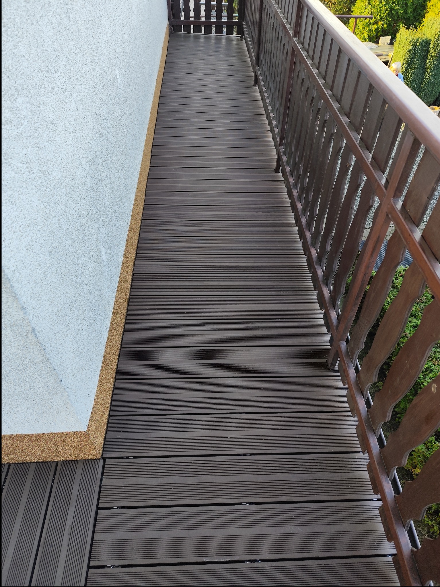 Długi balkon wyłożony ciemnobrązowymi panelami kompozytowymi, z drewnianą balustradą i widoczną strukturą ściany budynku z dekoracyjnym wykończeniem.