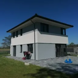 Dwukondygnacyjny dom z szarą elewacją, białą górną częścią, czarnymi ramami okiennymi i kostką brukową na podwórku, z niebieskim konikiem na biegunach.