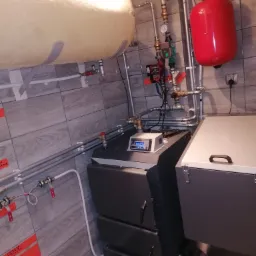 Nowoczesna kotłownia z piecem na paliwo stałe, zasobnikiem ciepłej wody i czerwonym naczyniem wzbiorczym, instalacja z rur miedzianych, szara płytka imitująca drewno na ścianie, widoczne elementy...