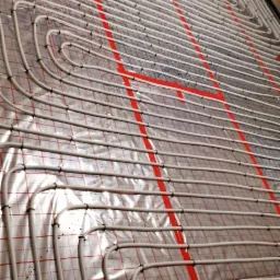 Białe rurki ogrzewania podłogowego ułożone na folii z czerwoną siatką, mocowane czarnymi klipsami.