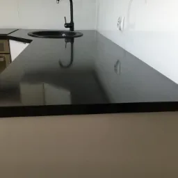 Czarny, błyszczący blat kuchenny z okrągłym, wpuszczanym zlewem i czarną baterią, zamontowany na białych szafkach, z piekarnikiem widocznym w tle.