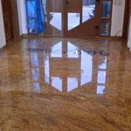 Błyszcząca, polerowana posadzka z kamienia o ciepłej, złoto-brązowej barwie z wyraźnym, falistym wzorem, z napisem 'Villa Roza' wkomponowanym w powierzchnię, odbijająca światło z drzwi wejściowych.