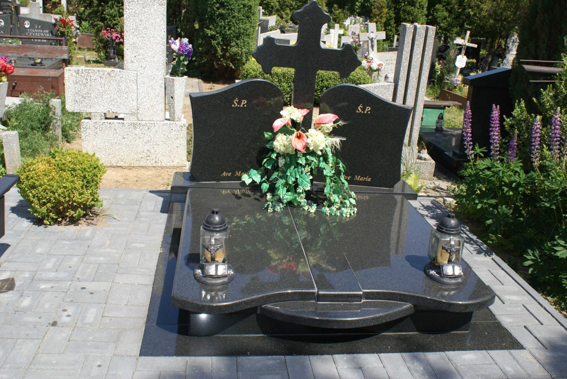 Czarny, podwójny nagrobek z krzyżem, ozdobiony kwiatami i lampionami, na cmentarzu z kostki brukowej.