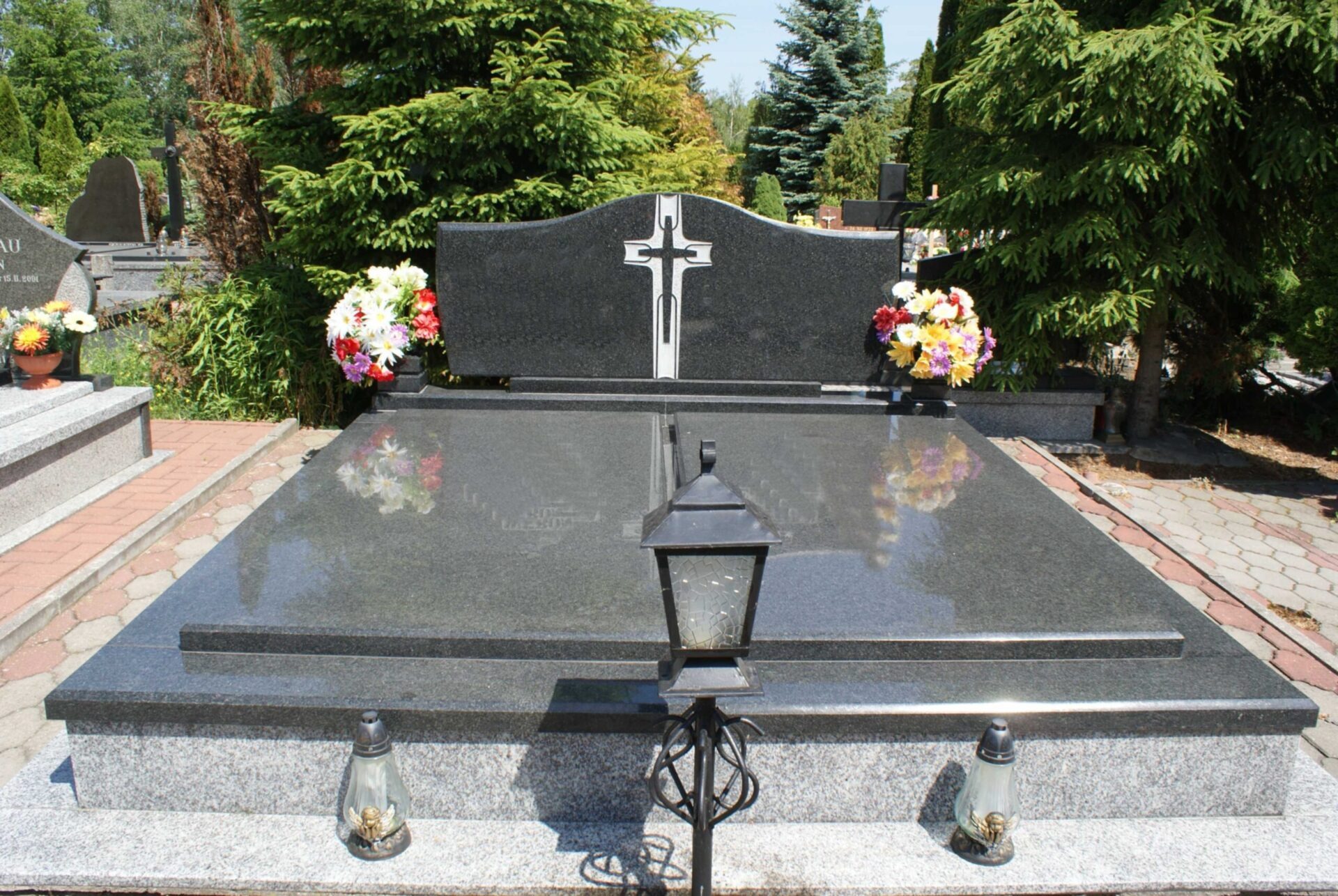 Czarny, podwójny nagrobek z granitu z białym krzyżem, ozdobiony kolorowymi kwiatami i lampionami, na tle zieleni cmentarza.
