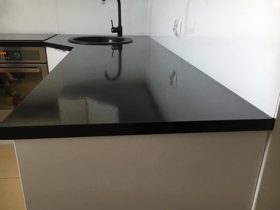 Czarny, błyszczący blat kuchenny z okrągłym, wpuszczanym zlewem i czarną baterią, zamontowany na białych szafkach, z piekarnikiem widocznym w tle.