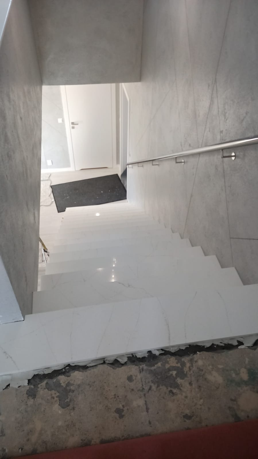 Wnętrze klatki schodowej z białymi stopniami imitującymi marmur i szarą ścianą z metalową poręczą, widoczne niedociągnięcia wykończeniowe przy podłodze.