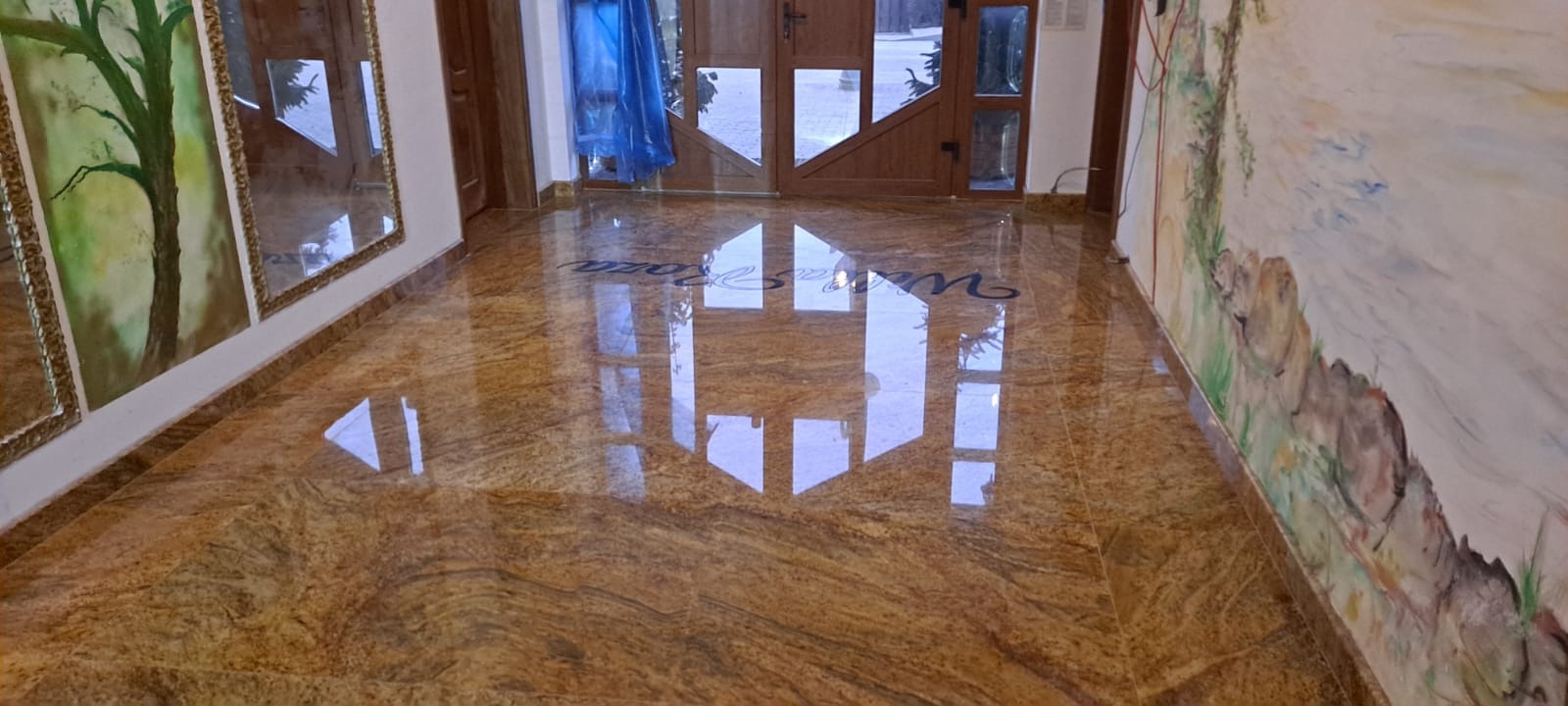 Błyszcząca, polerowana posadzka z kamienia o ciepłej, złoto-brązowej barwie z wyraźnym, falistym wzorem, z napisem 'Villa Roza' wkomponowanym w powierzchnię, odbijająca światło z drzwi wejściowych.