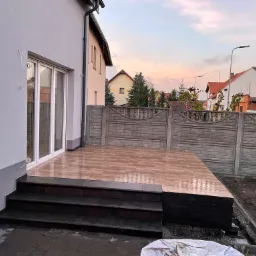 Nowo wyłożony, brązowy taras z płytkami ceramicznymi, z ciemnymi schodami prowadzącymi do białych, przeszklonych drzwi balkonowych, na tle szarego domu i betonowego ogrodzenia.