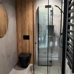 Nowoczesna łazienka z kabiną prysznicową typu walk-in z czarnymi profilami, podwieszaną toaletą w czarnym kolorze, drewnianą okładziną ścienną i szarą, betonową fakturą na ścianach.