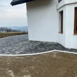 Ułożona kostka brukowa w odcieniach szarości i czerni, z jasną obwódką, przylegająca do białego budynku z brązowymi oknami, na tle krajobrazu z domami i wzgórzami.