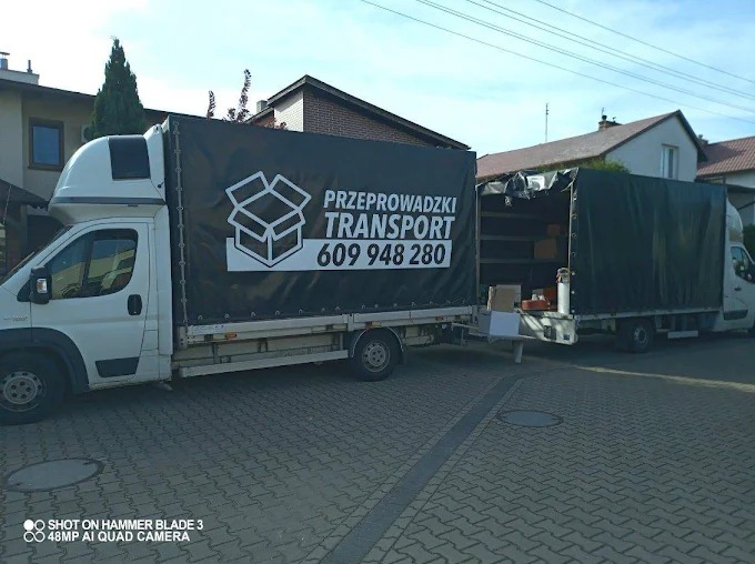Dwa busy transportowe z otwartą przestrzenią ładunkową, częściowo załadowane kartonami, na tle domów jednorodzinnych. Na plandece busa widnieje napis: Przeprowadzki Transport.