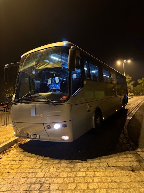 Szary autobus Nova z otwartymi drzwiami, zaparkowany nocą na brukowanej ulicy pod latarnią. Widoczne oświetlenie pojazdu i wnętrze z niebieskimi zagłówkami.