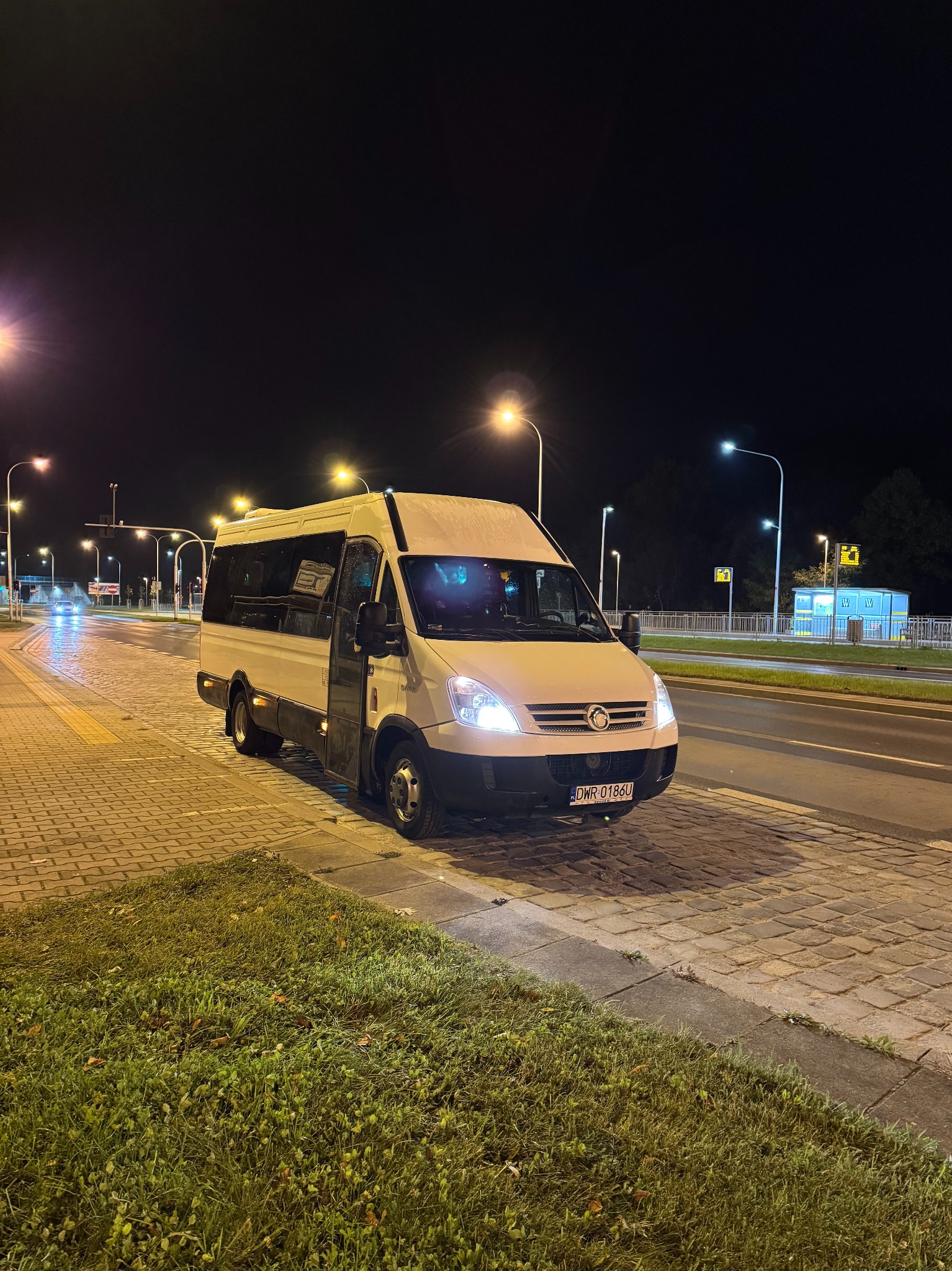 Biały bus z otwartymi drzwiami stoi nocą na ulicy, oświetlony przez latarnie. Widoczna tablica rejestracyjna i fragment chodnika z kostki brukowej.