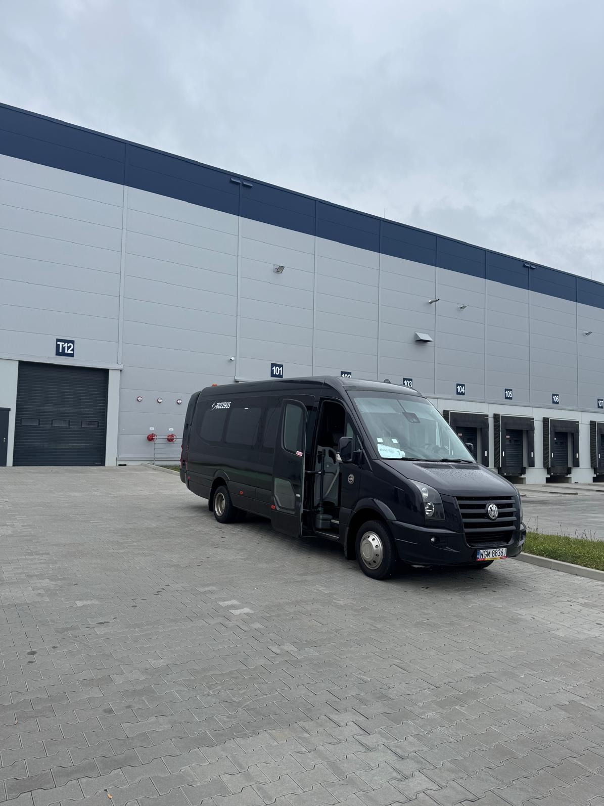 Czarny bus z otwartymi drzwiami na tle magazynu z numerami bram. Widoczny fragment chodnika z kostki brukowej i tabliczka z numerem T12.