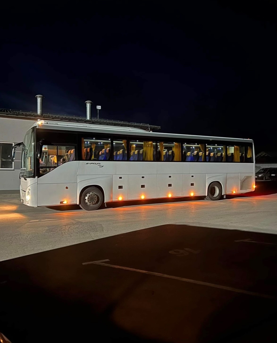 Biały autokar z włączonymi światłami postojowymi stoi na parkingu nocą. Widoczne zasłony w oknach i rząd siedzeń. Nadwozie z napisem 'EUROPA BUS'.