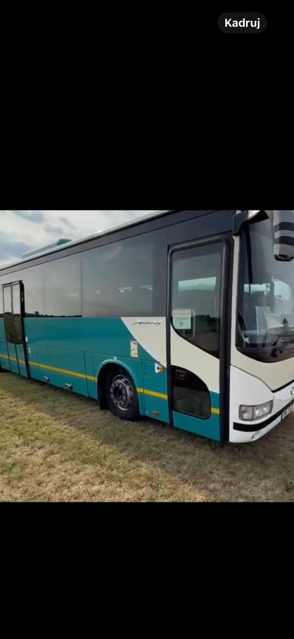 Turkusowo-biały bus na trawiastym terenie, widok z boku, lekko pochmurne niebo w tle. Pojazd z dużymi oknami i czarnym dachem.