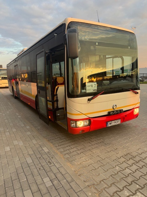 Autobus miejski z otwartymi drzwiami na przystanku, widoczne odbicie zachodzącego słońca w szybach, tablica rejestracyjna DWR 9439T.