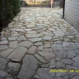 Ścieżka z nieregularnych płyt kamiennych w ogrodzie, z niskim oświetleniem punktowym wbudowanym w nawierzchnię, prowadząca do bramy z metaloplastyki i budynku z jasnej cegły.