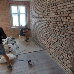 MagMarBud - Mężczyzna wierci w ceglanej ścianie w trakcie prac remontowych, widoczne narzędzia i materiały budowlane, podłoga drewniana częściowo zakryta folią ochronną.